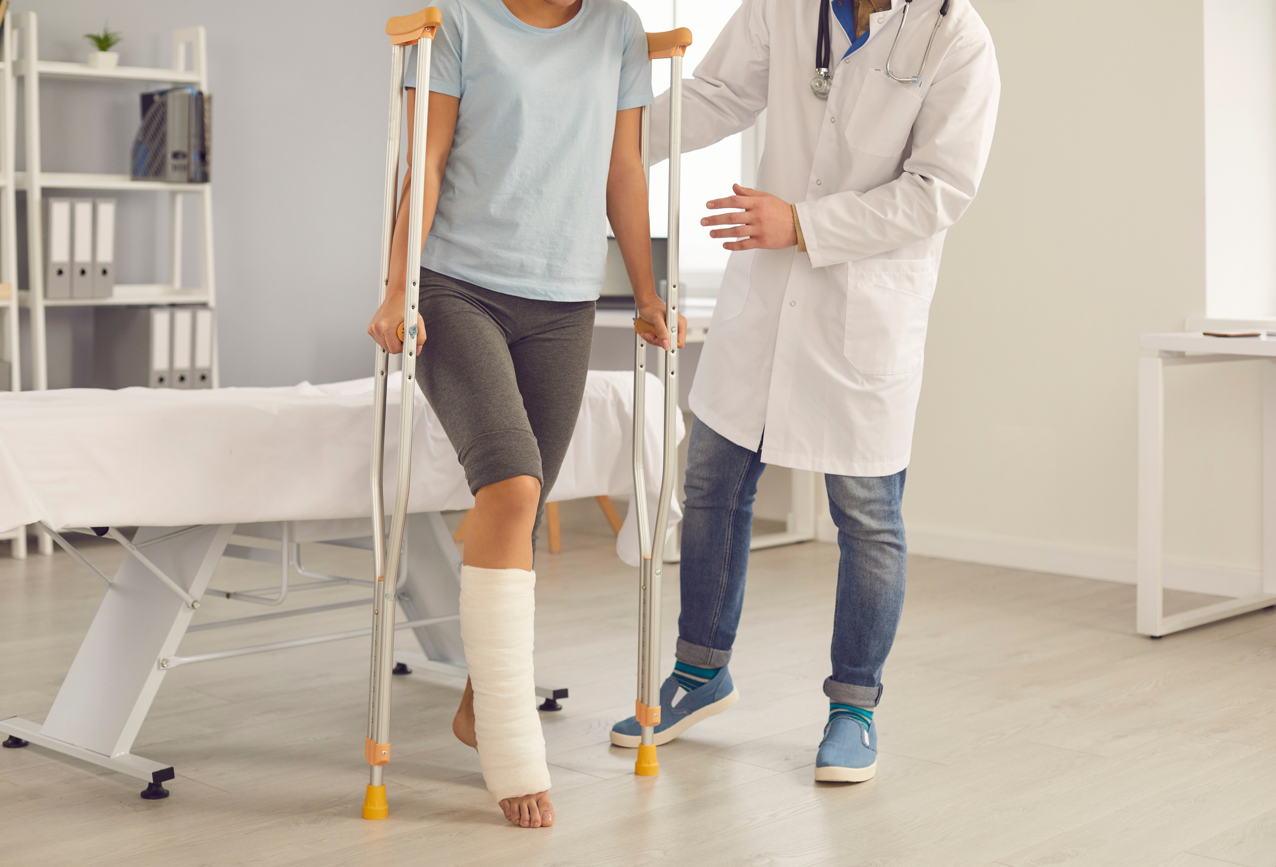 Doctor Supports a Patient with a Broken Leg While Walking on Crutches in a Hospital Office.